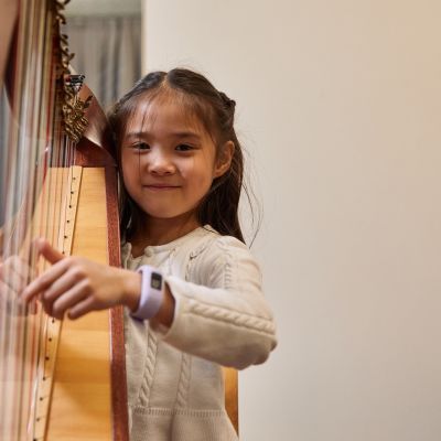 A girl with black hair and wearing a white knitted dress smiles at the camera whilst playing a harp